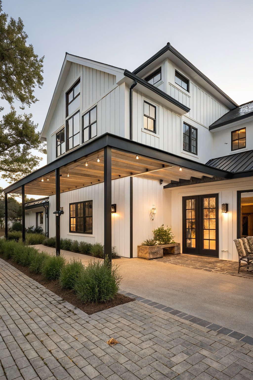 Corner view of a two-story white board-and-batten farmhouse with black window frames, black steel covered porch supported by posts, string lights, black double doors entry, potted plant, Adirondack chair, and brick paver path edged by grasses.