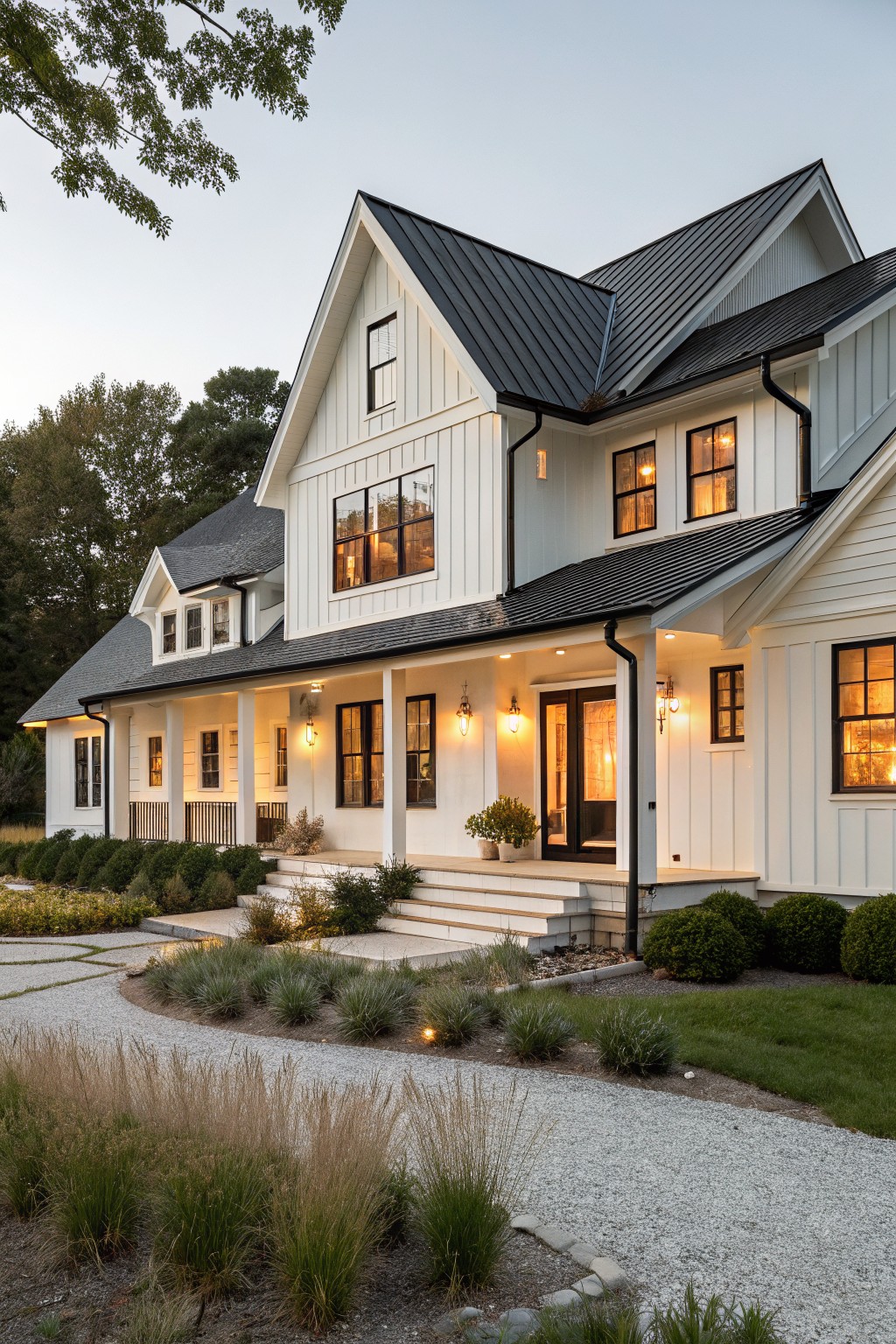 Two-story white board-and-batten farmhouse exterior with black standing-seam metal roof, black window and door trim, wraparound front porch, steps to entry, gravel pathway, and ornamental grasses at dusk.