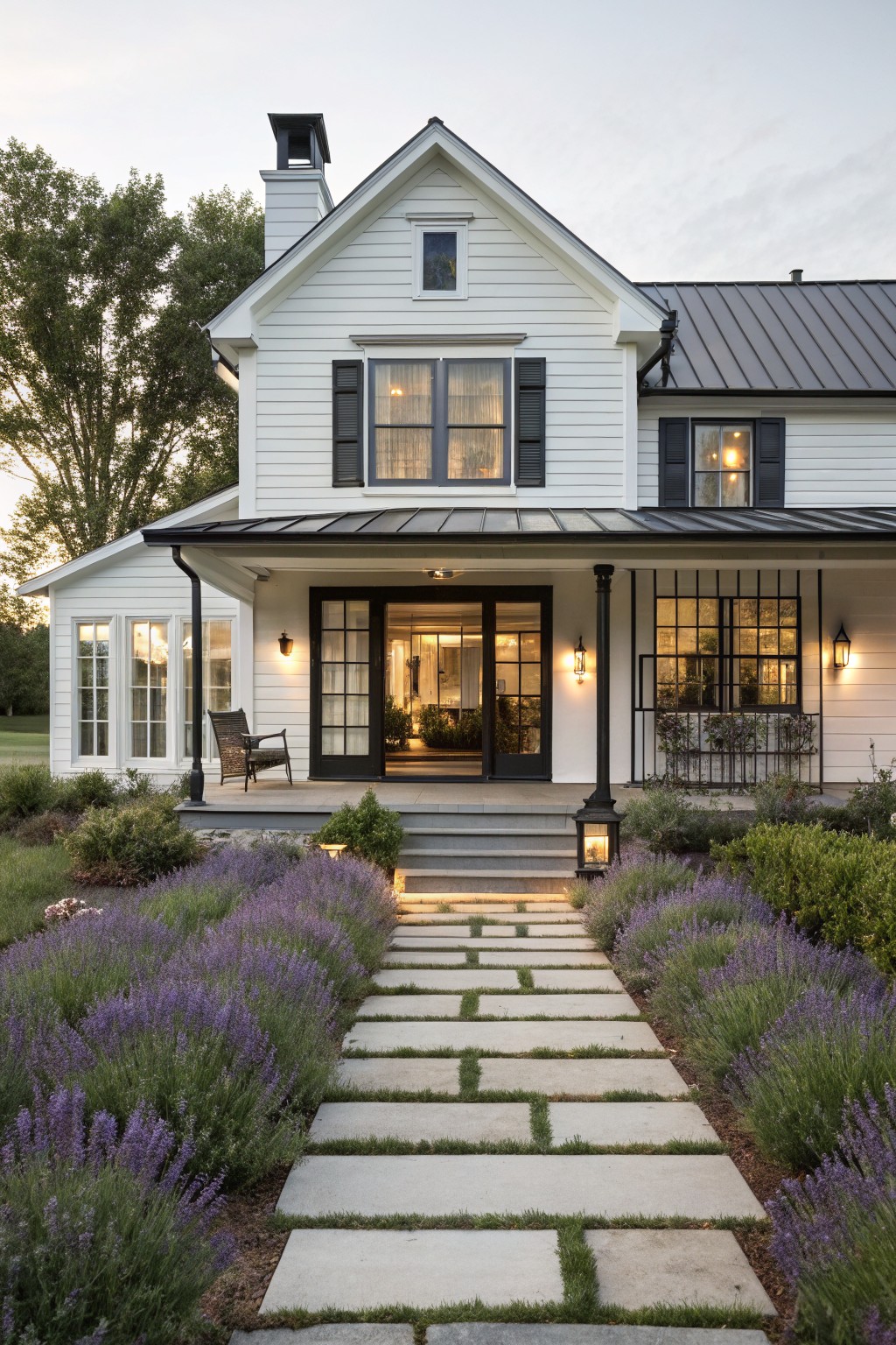 White clapboard farmhouse with black shutters, black metal porch roof and columns, black-framed glass entry doors, dark metal main roof, lanterns, and stone pathway bordered by lavender plants leading to the front steps.