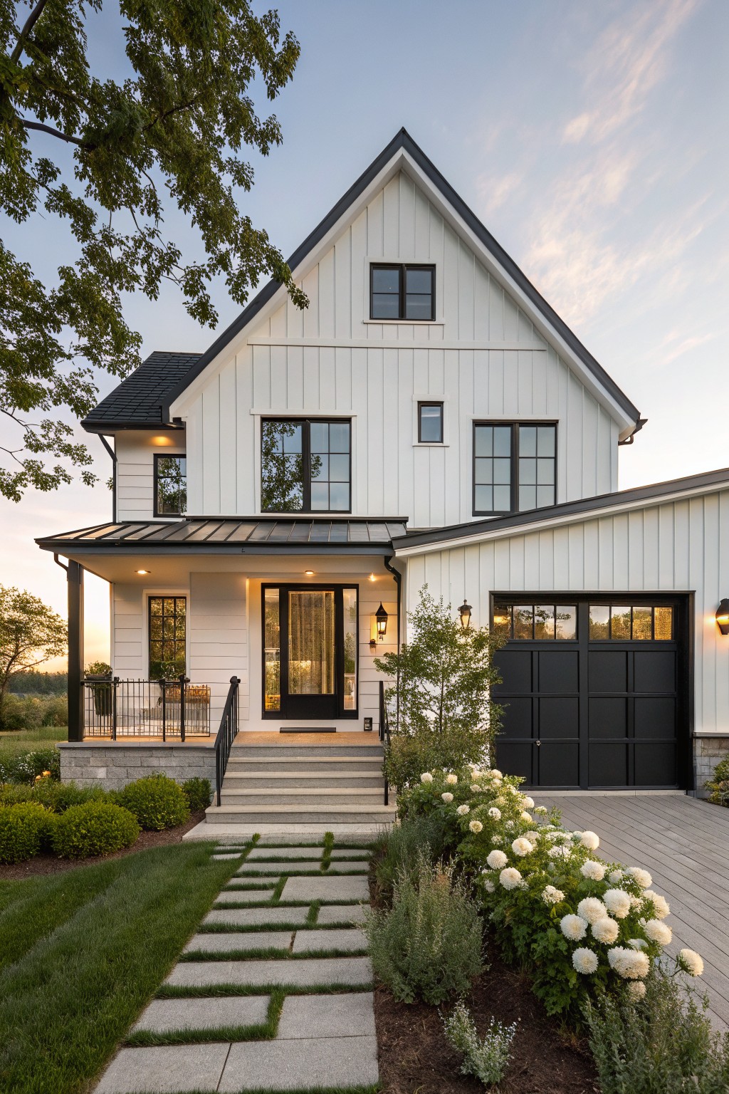 A two-story white board-and-batten farmhouse with black-framed windows, front door, and garage door, featuring a covered porch, stone steps, paver pathway, and hydrangea plantings in the front yard at dusk.