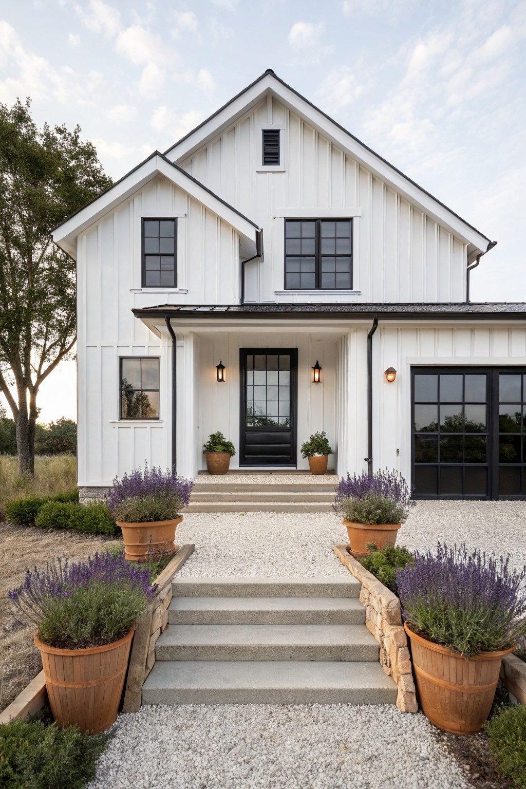 White board-and-batten sided two-story modern farmhouse house exterior featuring black-trimmed windows, front door with sidelights, and garage door under a covered porch, with concrete steps flanked by stone retaining walls and large terracotta pots of lavender plants, gravel driveway and landscaping.