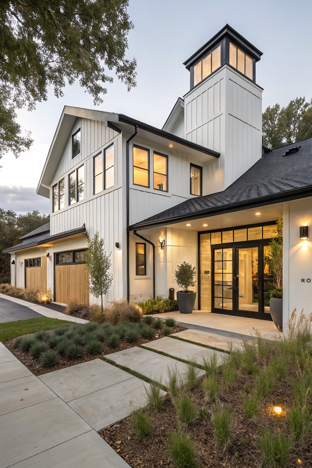 White modern farmhouse exterior at dusk with tall cupola tower, black trim on windows and doors, wood garage doors, glass entry, and low native plant landscaping along concrete path and driveway.