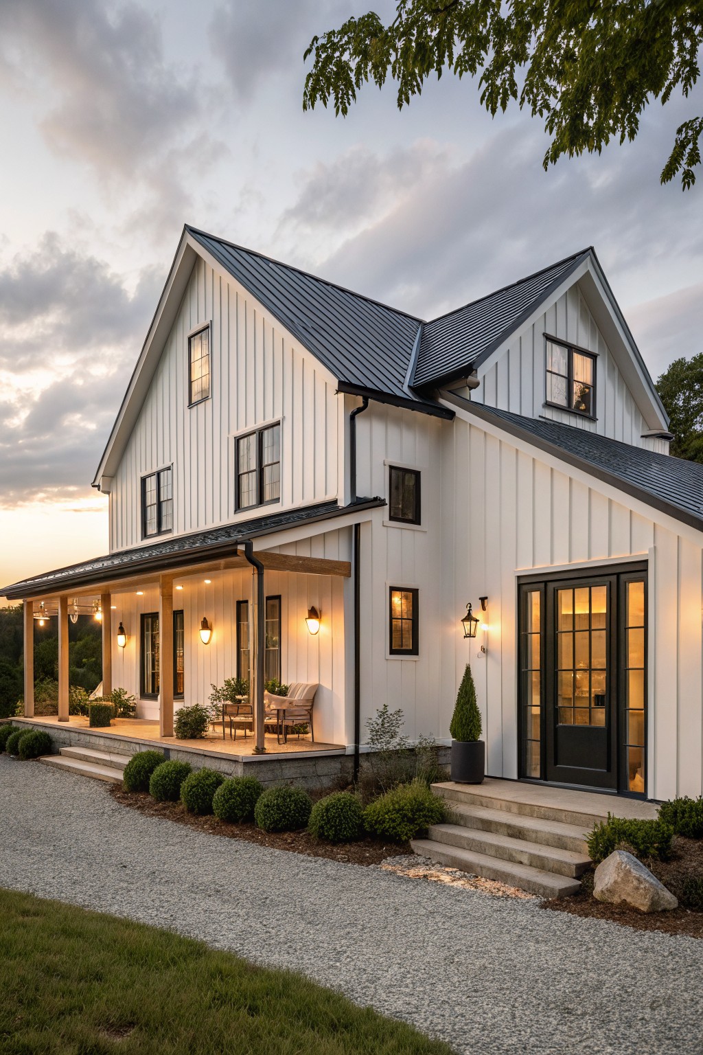 Two-story white board-and-batten farmhouse exterior with black metal gable roof, black-trimmed windows and doors, covered front porch with chairs and plants, gravel driveway, boxwoods, and landscape lighting at dusk.