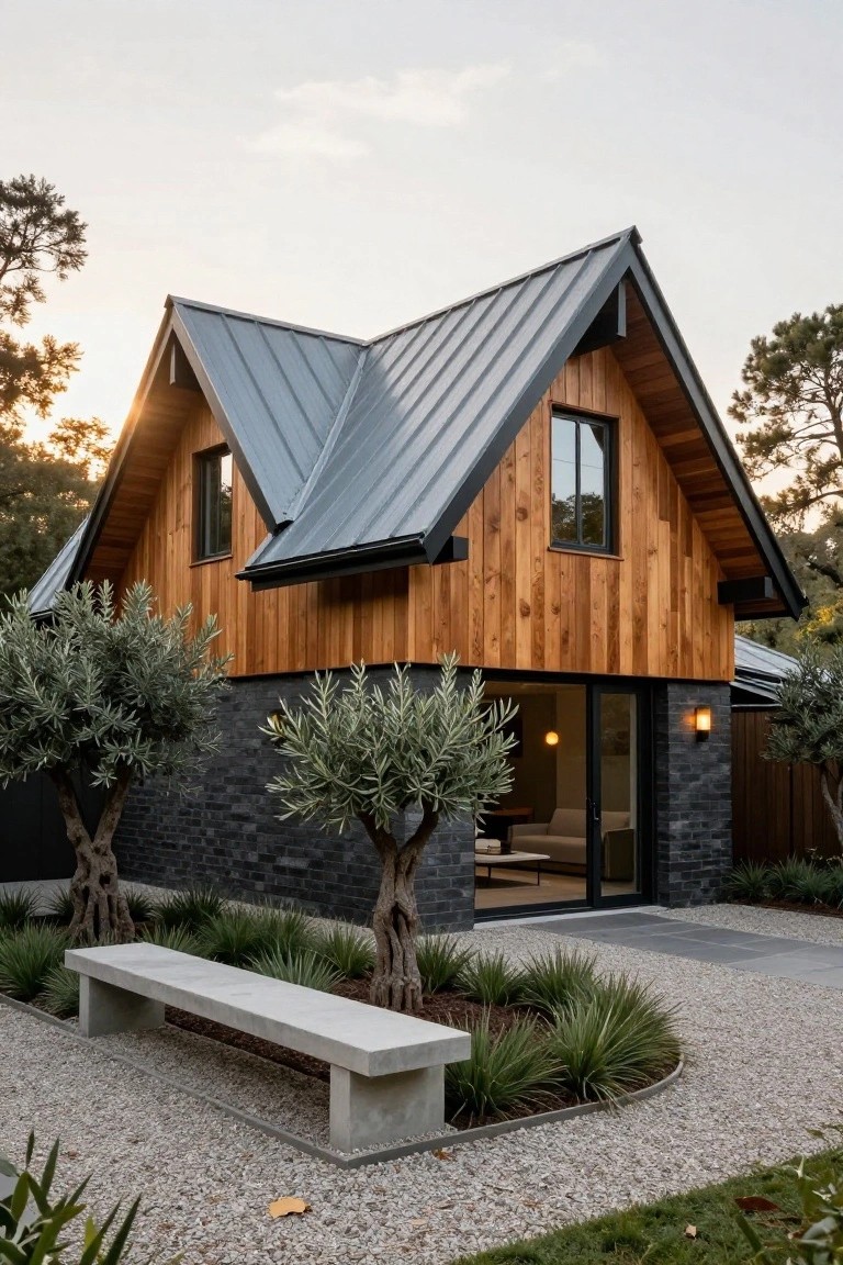 Modern house exterior with steeply angled gray metal roof, vertical orange-toned wood cladding, black brick base, glass entry doors, olive trees, concrete bench, and gravel path.