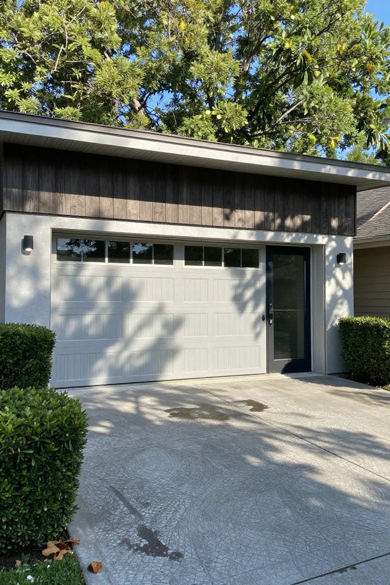 White overhead garage door with dark horizontal wood siding above it on a house exterior, black side door, wall lights, boxwood shrubs on both sides, and concrete driveway with tire marks.