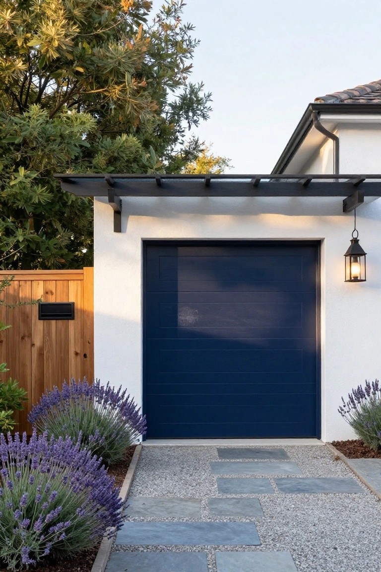 Navy blue garage door under a black wood pergola on a white stucco wall, flanked by lavender plants, a wooden fence, gravel ground cover, and stone pavers leading to the door.