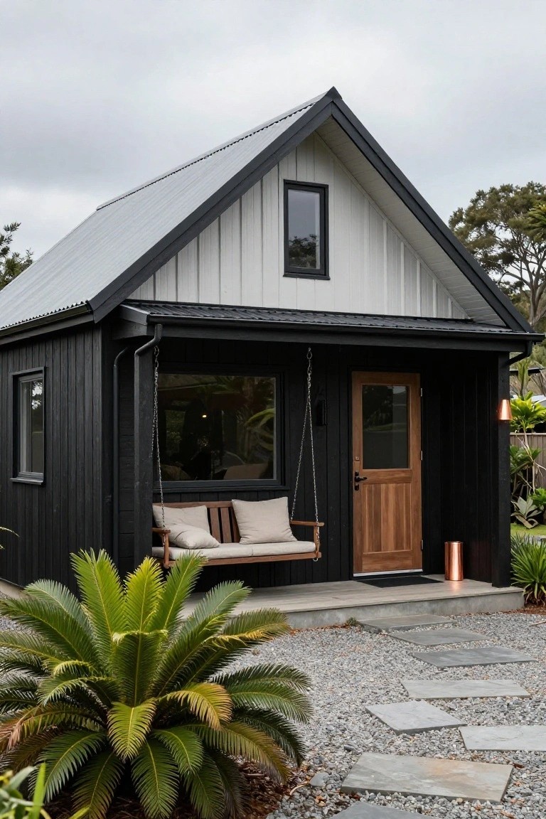 Small gabled cabin with black shiplap siding on lower walls, white cladding on upper gable, covered porch with hanging bench swing, wooden entry door, copper light, and potted plants on gravel yard with stone path.