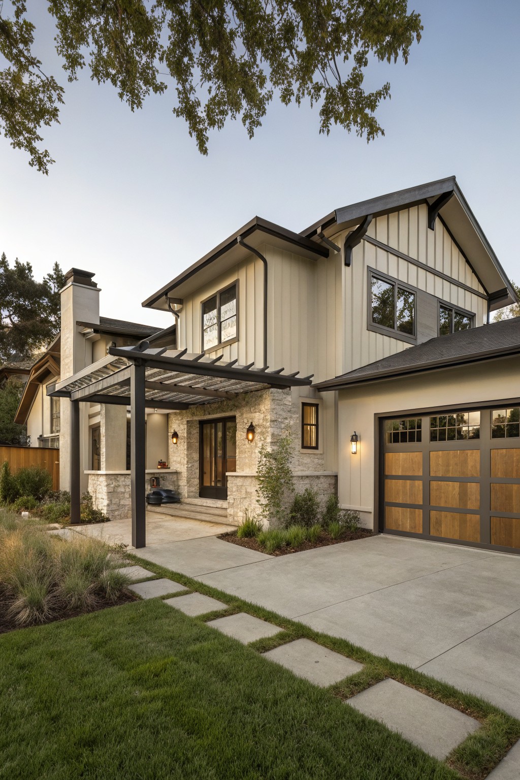 Modern farmhouse house exterior with light beige vertical siding, black trim and roof, stone entry porch under a metal pergola, wood garage door, and surrounding lawn and plants.