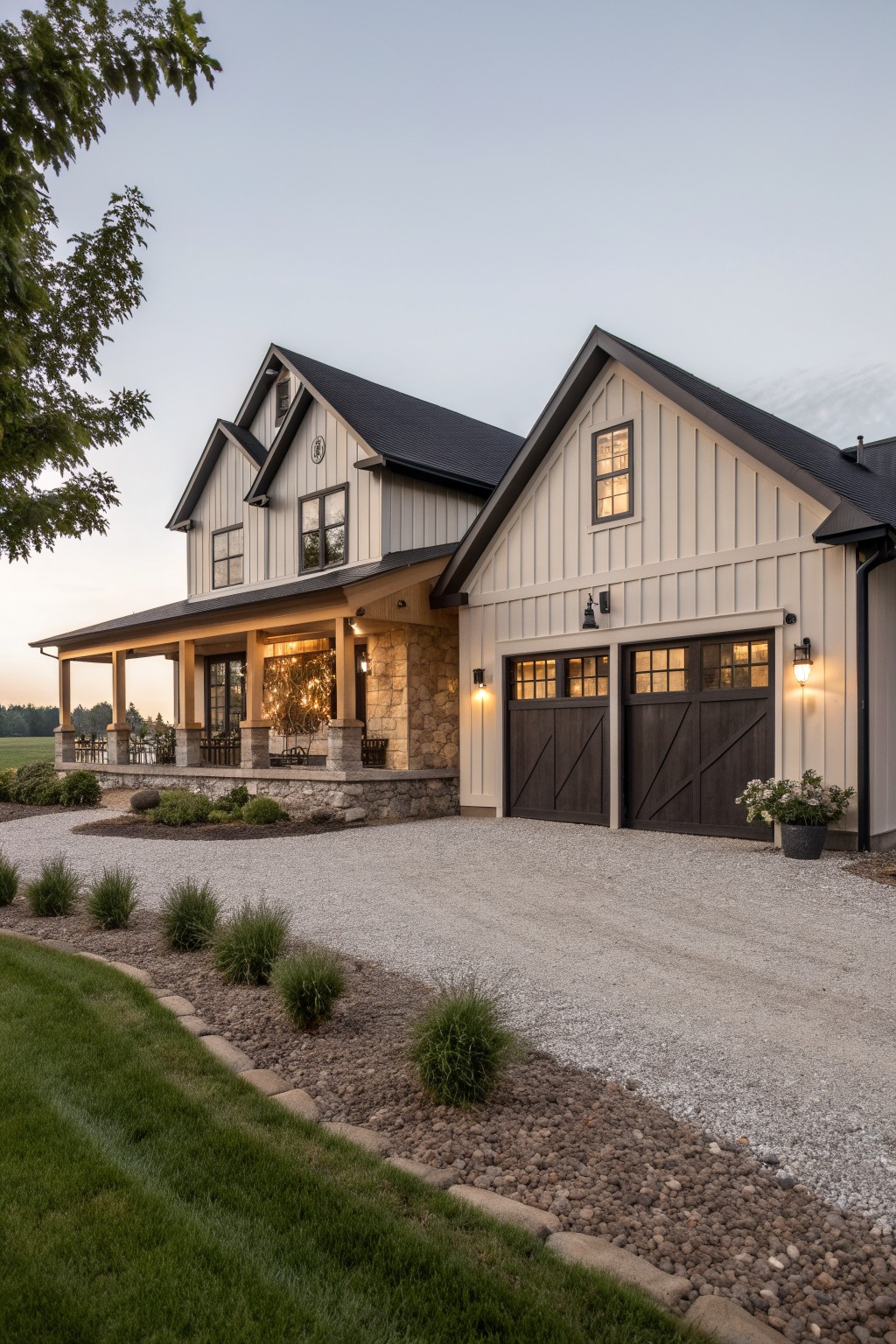 Modern farmhouse house with light gray board-and-batten siding, black asphalt shingle roof, dark wood double garage doors, covered front porch with stone base, gravel driveway, and low landscaping along the edges.