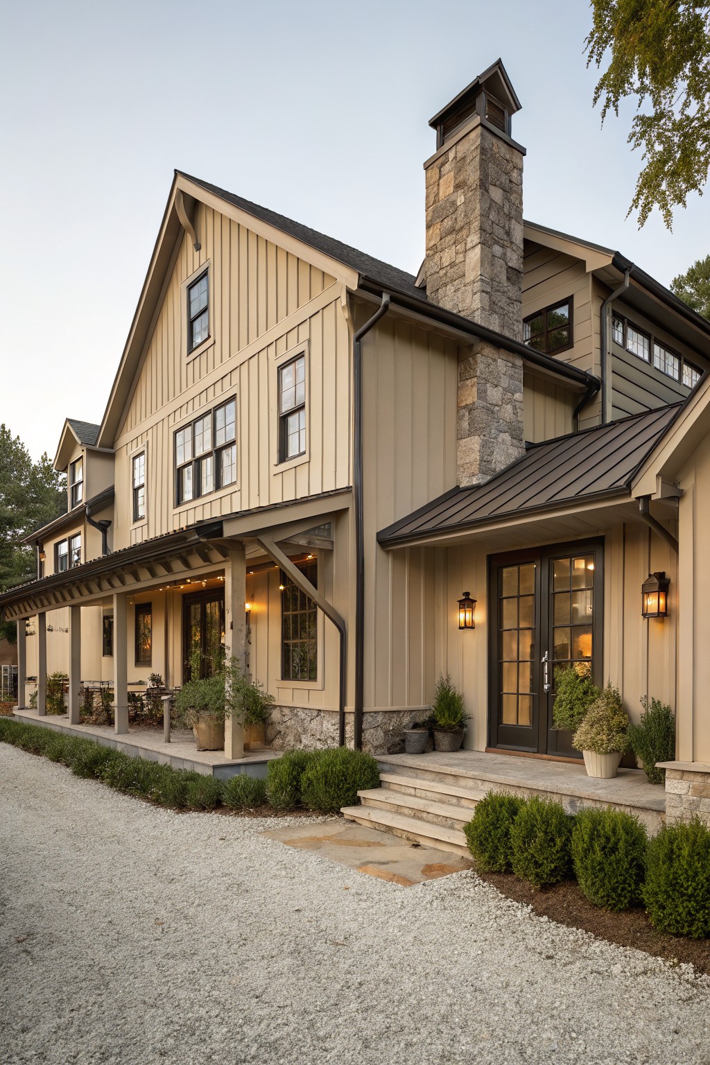 Side view of a modern farmhouse exterior with light beige board-and-batten siding, tall stone chimney, metal roof sections, wraparound porch, lanterns by glass doors, gravel driveway, and low shrubs.