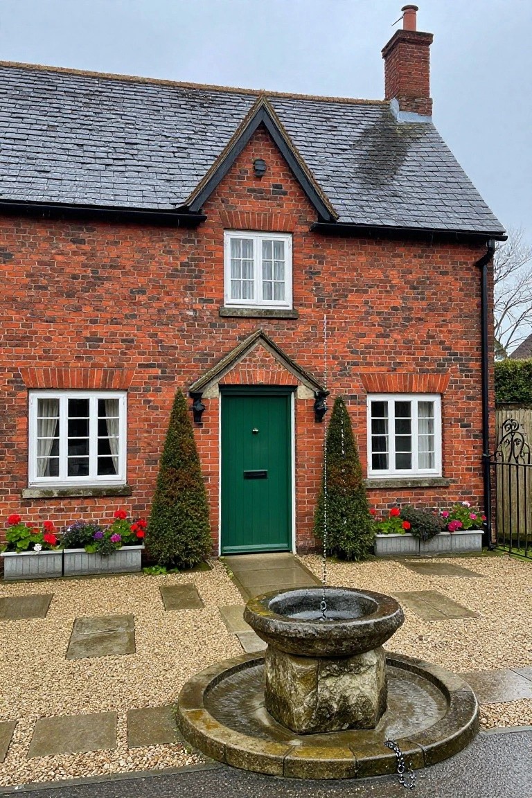 Red brick cottage with black timber framing, slate roof, white windows, green front door flanked by conical topiaries and flower boxes, stone fountain in gravel front yard under overcast sky.