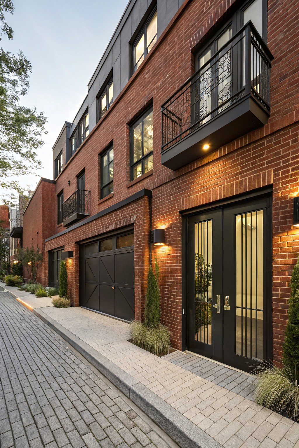 Multi-story red brick townhouse exterior with black garage doors, double entry doors, balconies, and landscaping along a cobblestone pathway.