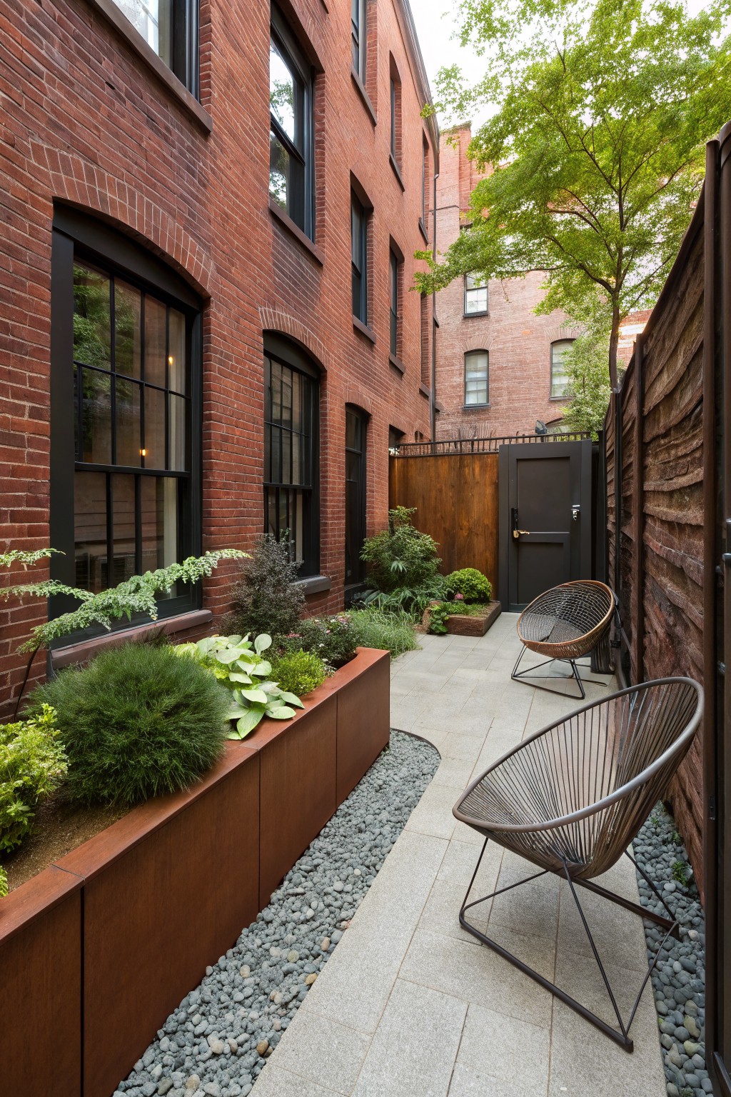 Narrow vertical view of a brick-walled courtyard with tall red brick buildings on both sides, a dark wooden gate on a fence, raised rectangular corten steel planter filled with greenery along one wall, stone paver path edged with gravel, and two black woven wireframe lounge chairs positioned near the end.