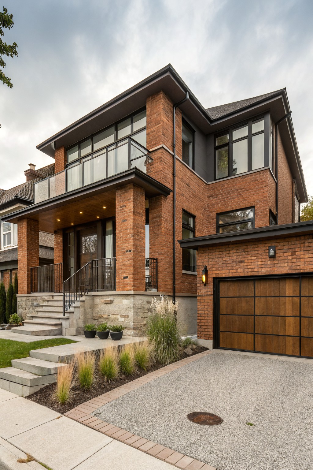 Two-story red brick house exterior showing front porch with wood ceiling and lighting, glass-railed balcony above, dark-framed windows, wooden garage door, stone steps, and ornamental grasses in the yard.