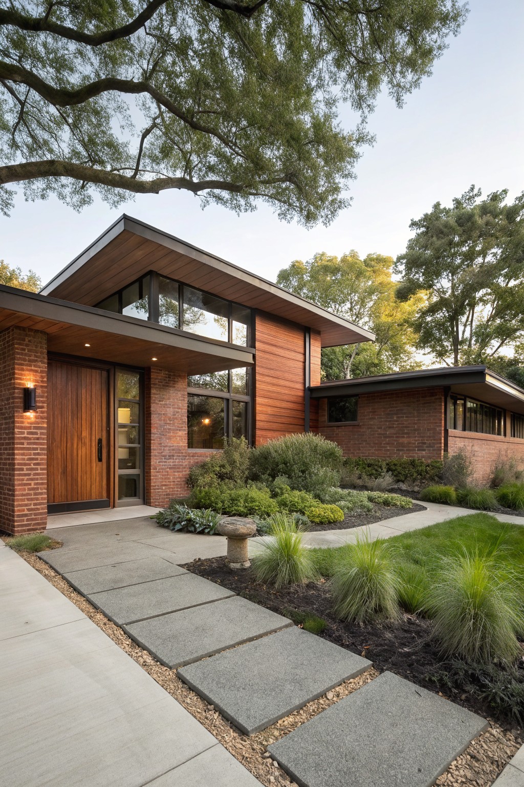 Exterior of a single-story red brick house with cedar wood siding accents, a wide overhanging flat roof above a vertical wood entry door, glass windows, and a concrete paver path through low landscaping.