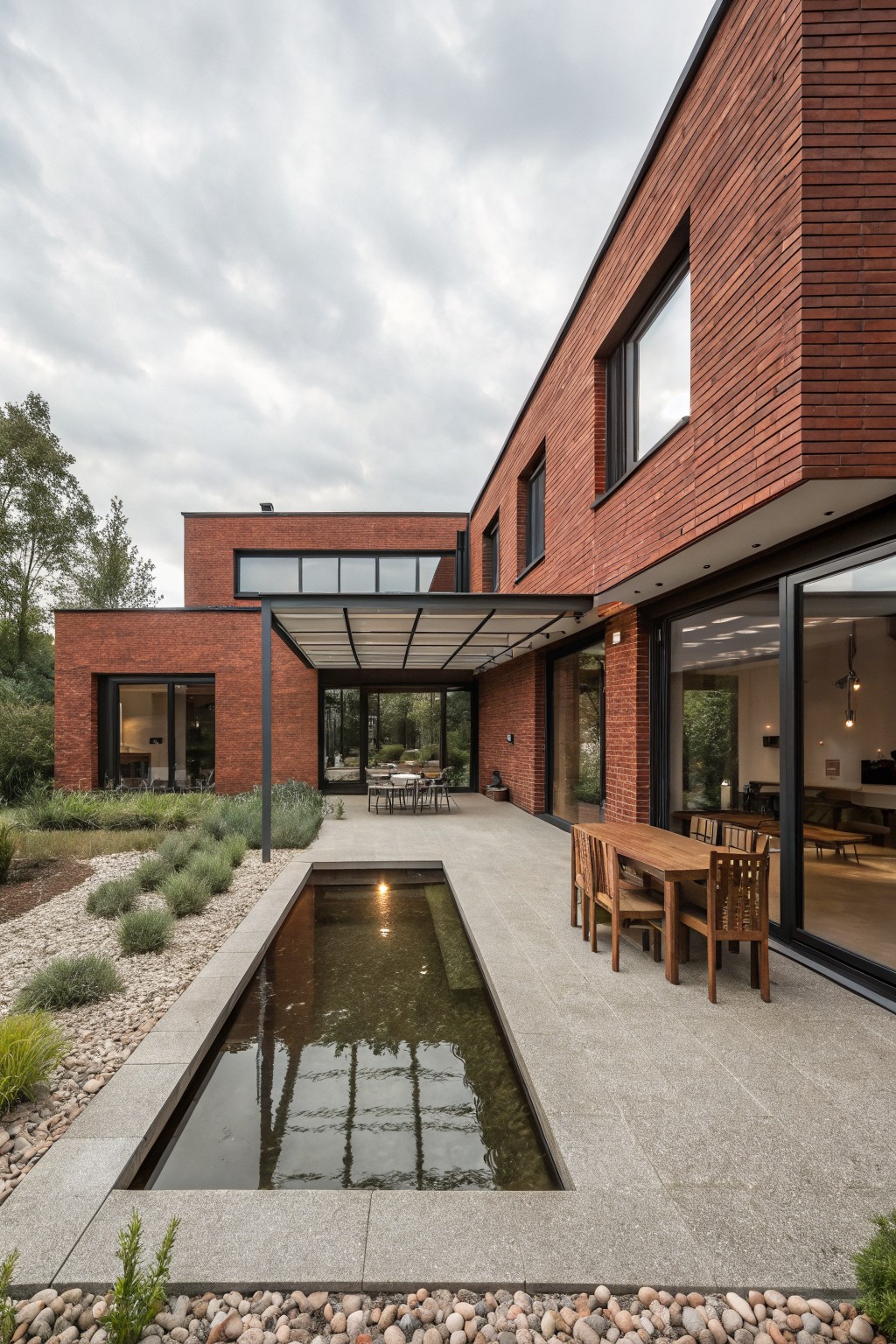 Side exterior of red brick house with dark-framed windows and glass doors opening to concrete terrace under white metal pergola, wooden dining table with chairs, narrow reflecting pool, gravel ground cover, and low plants.