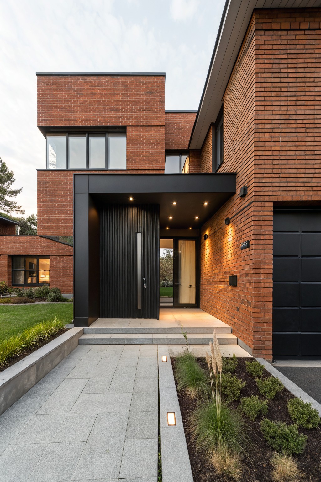 Red brick house exterior featuring a black cantilevered entry canopy with vertical slat door, concrete steps and pathway, black garage door, and low plantings along the front.