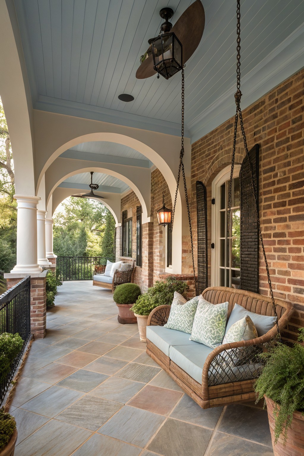 Covered porch with red brick walls, white arches and columns, blue painted plank ceiling, hanging wicker swing with cushions, potted plants, ceiling fans, lanterns, and stone tile floor.