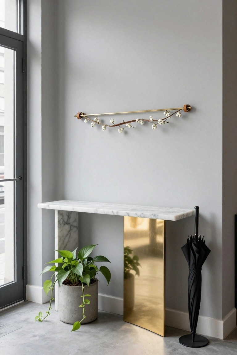 Light gray interior wall with a horizontal gold rod mounted on wooden brackets holding a curving branch with clusters of white berries, above a white marble console table on a tall brass pedestal base, a potted trailing green plant in concrete pot, and black umbrella stand next to a dark metal and glass door.