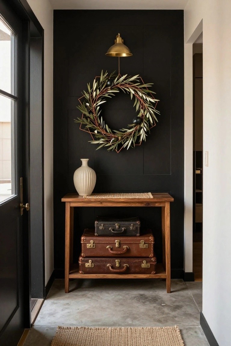 Modern interior entryway with black accent wall displaying a geometric olive leaf wreath, gold sconce light above, white textured vase on wooden console table stacked with brown leather suitcases, black door, seagrass rug on concrete floor.