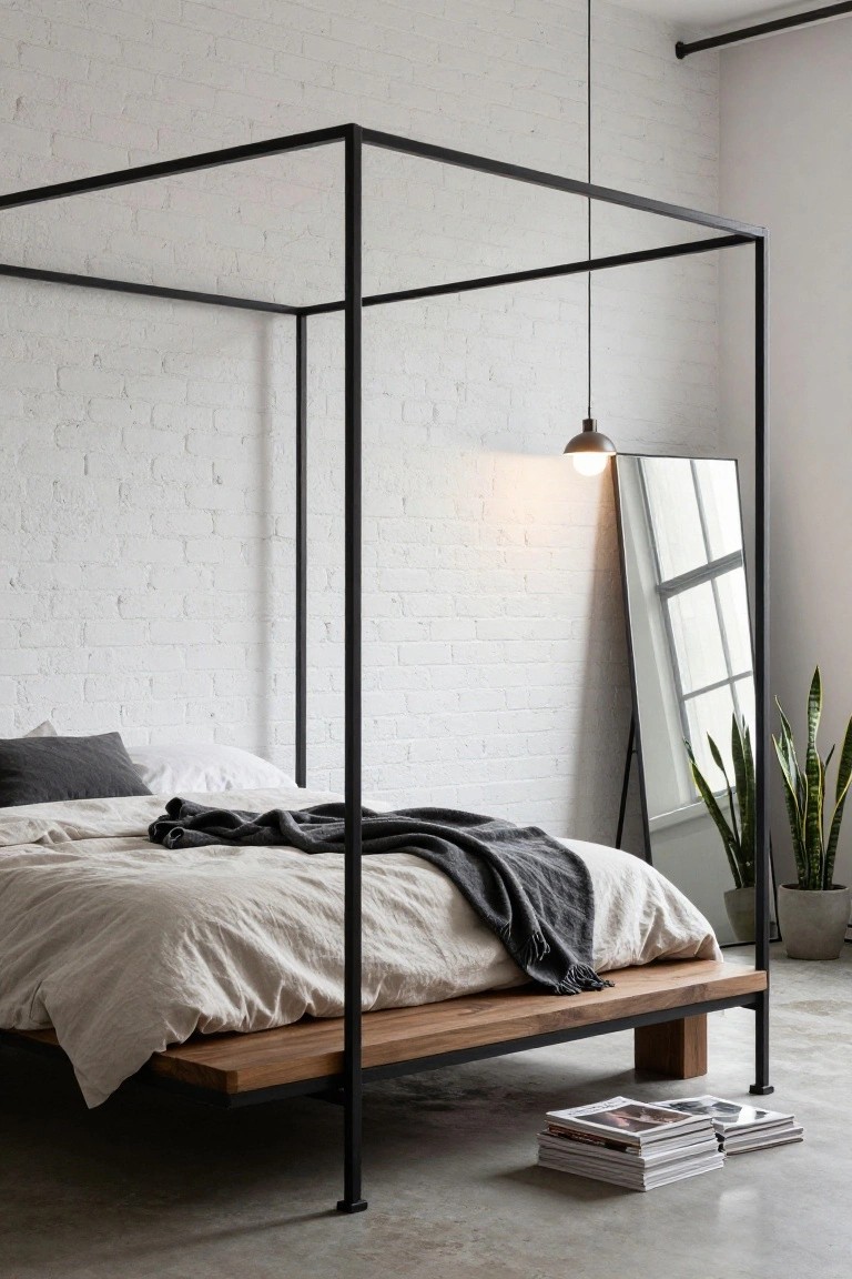 A minimalist bedroom with black metal canopy bed frame over low wooden platform bed, white brick walls, full-length mirror, potted snake plants, pendant light, and stacked magazines on concrete floor.