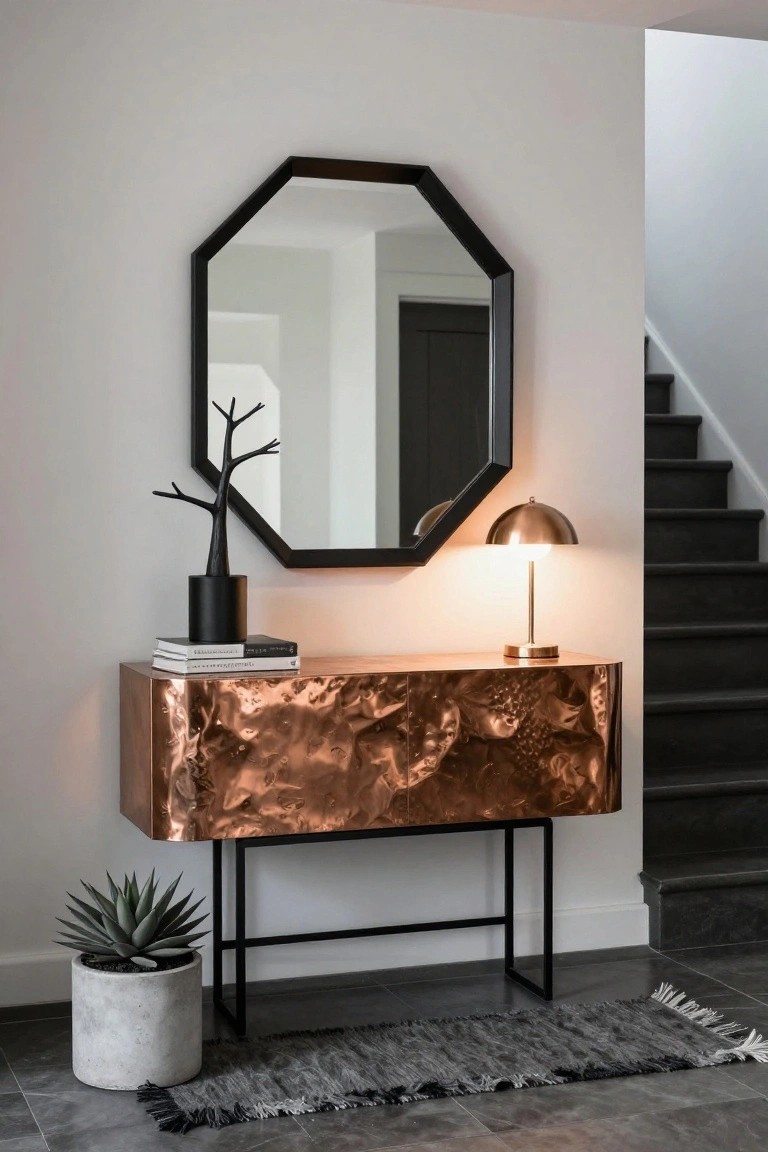 Modern entryway with black octagonal mirror over hammered copper console table on black metal legs, topped with black branch sculpture, books, brass lamp, and concrete pot with agave plant, beside dark wood staircase.