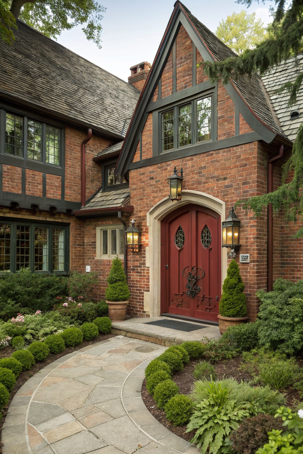 Red brick Tudor-style house exterior with dark green window frames and trim, arched red front door with wrought iron details, wall lanterns, stone pathway, and boxwood shrubs.
