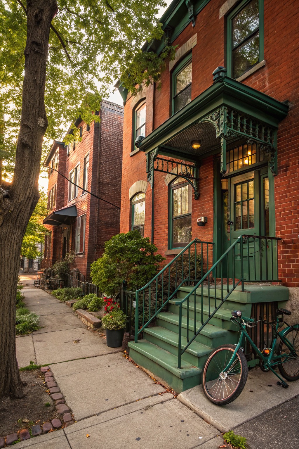 Red brick rowhouse exterior with green porch, arched entry door, metal railings, steps, and trim, plus small plants and a green bicycle at the base on a sidewalk lined with trees.