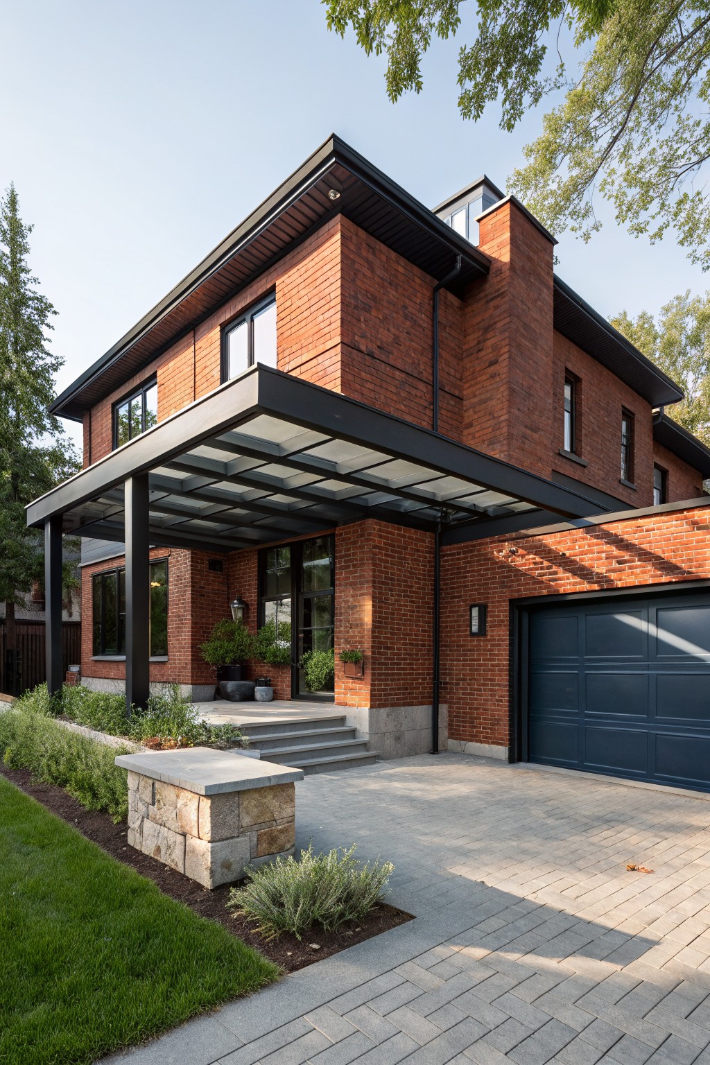 Two-story red brick house with black metal frames on windows and glass entry canopy, navy blue garage door, stone pillar by steps, low shrubs, and paved driveway.