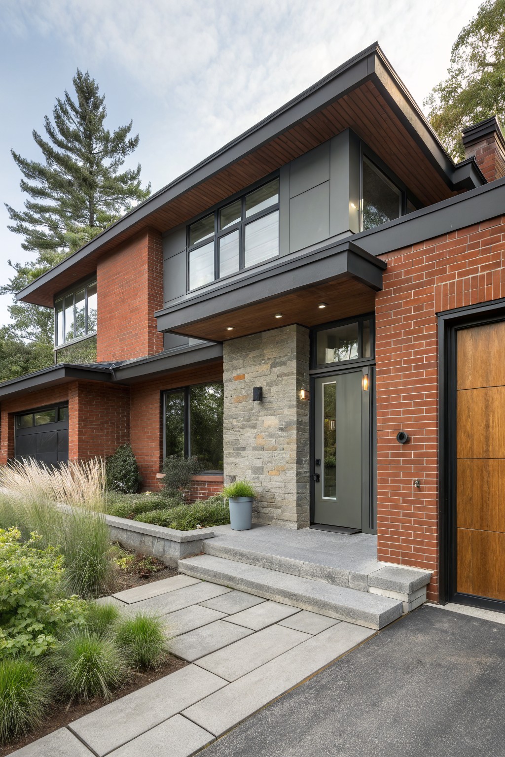 Two-story modern house exterior with red brick walls on the lower level, gray metal cladding on upper sections, wood soffits and accents, large windows, stone entry surround with gray door, attached wood garage door, concrete steps, and front landscaping with ornamental grasses and shrubs.