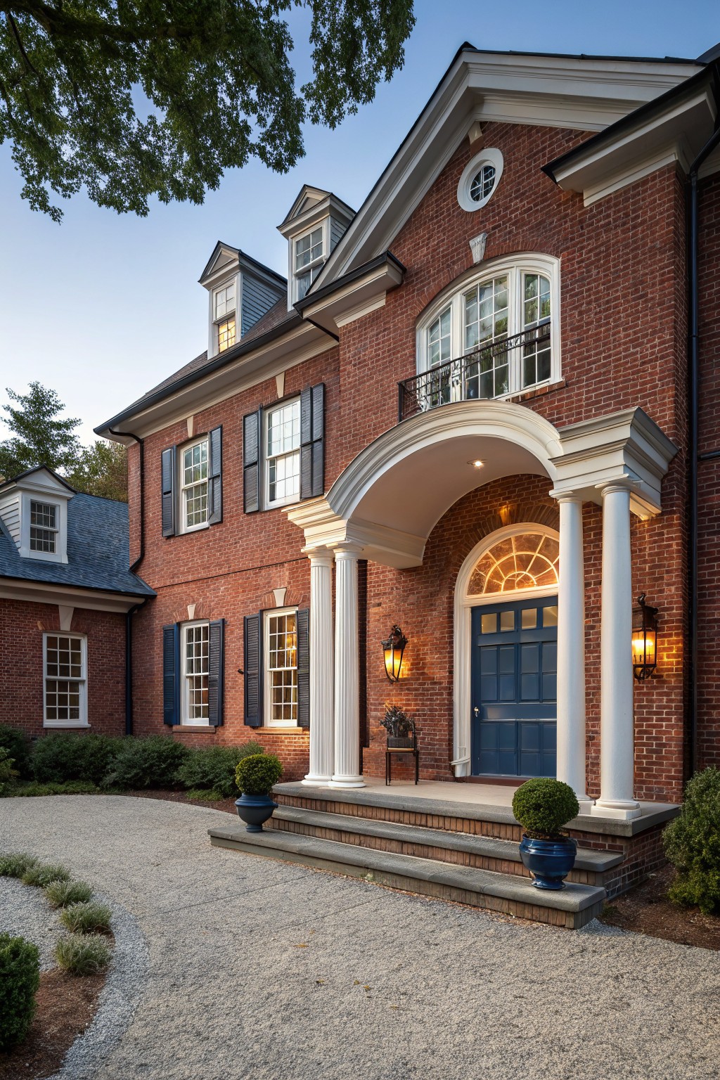 Two-story red brick house exterior featuring a navy blue front door under a white-columned portico, with black shutters, lanterns, topiary planters, and a curved gravel driveway edged in shrubs.
