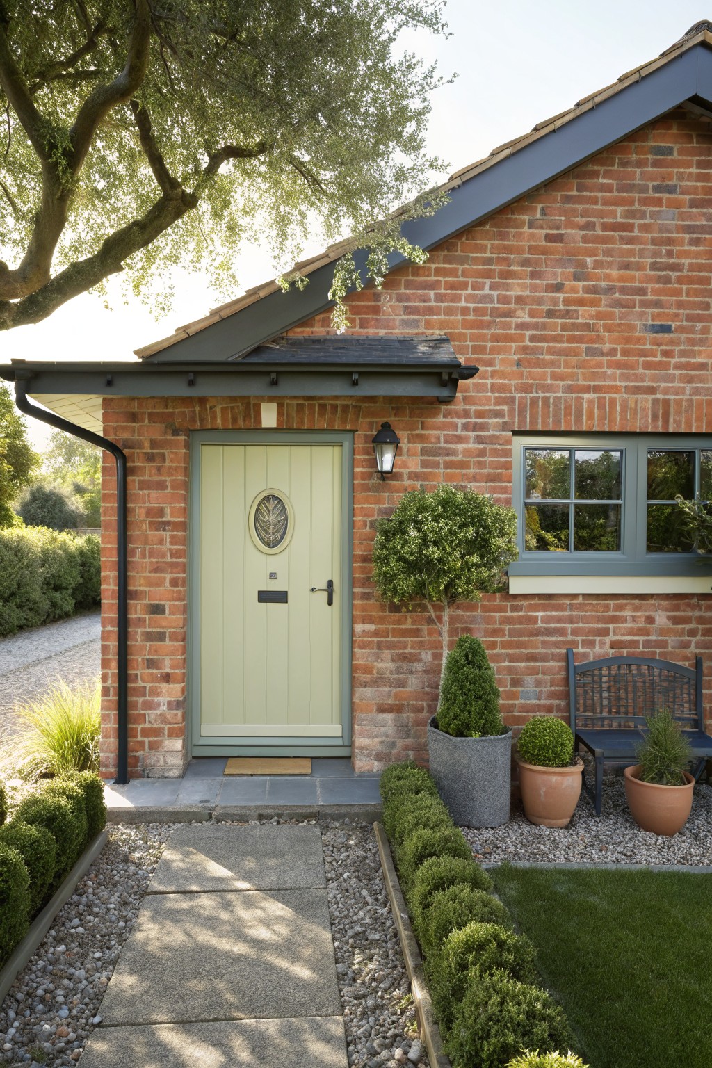 Red brick house exterior featuring a pale green paneled front door with oval window, flanked by potted topiary shrubs, a wooden bench, and a stone path edged with low hedges.