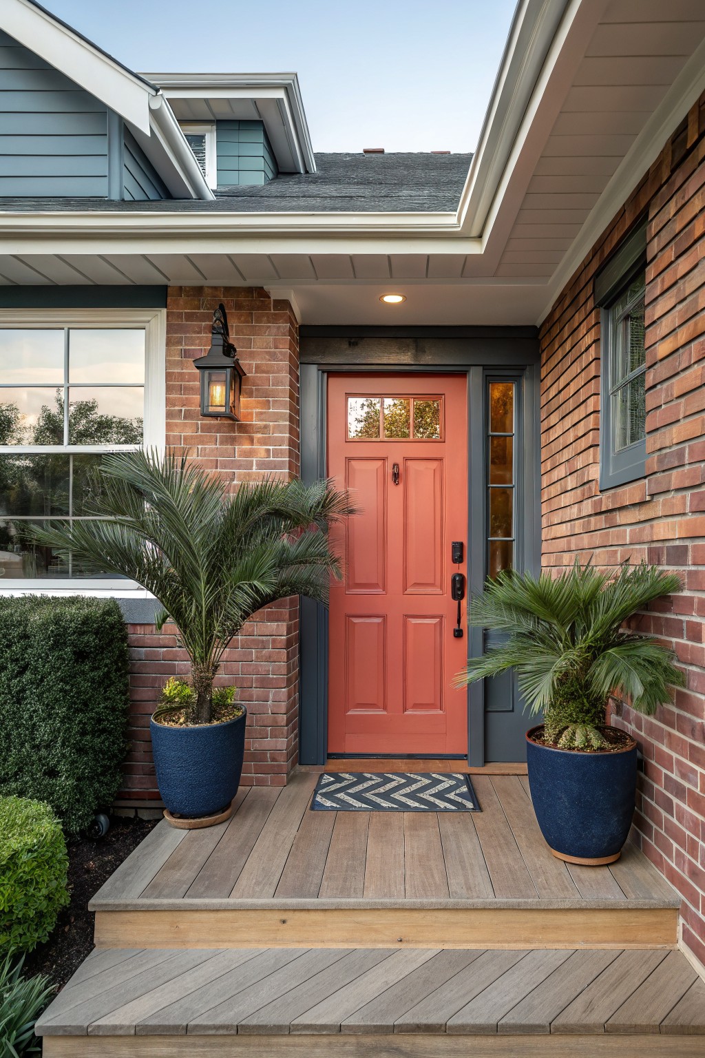 House exterior close-up showing red brick walls, blue siding, a bright orange-red paneled front door with sidelights, flanked by two blue pots with palm plants, a chevron welcome mat on a wooden porch step, and wall lanterns.