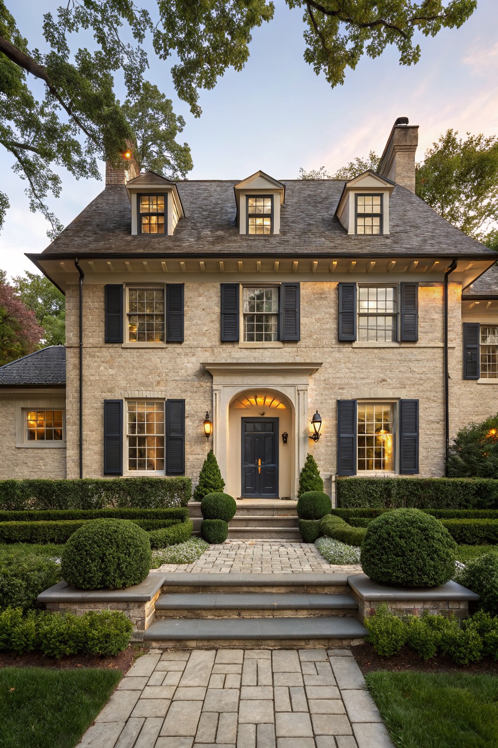 Two-story beige brick house with black shutters on multi-pane windows, a dark wood front door under an arched entry, flanked by boxwood shrubs, stone steps, and a bluestone pathway.