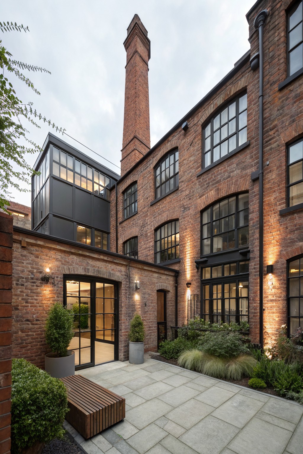Rear exterior of a red brick multi-story building with tall chimney, black-framed windows, modern glazed extension, paved courtyard terrace, wooden bench, potted topiary plants, and entrance doors.