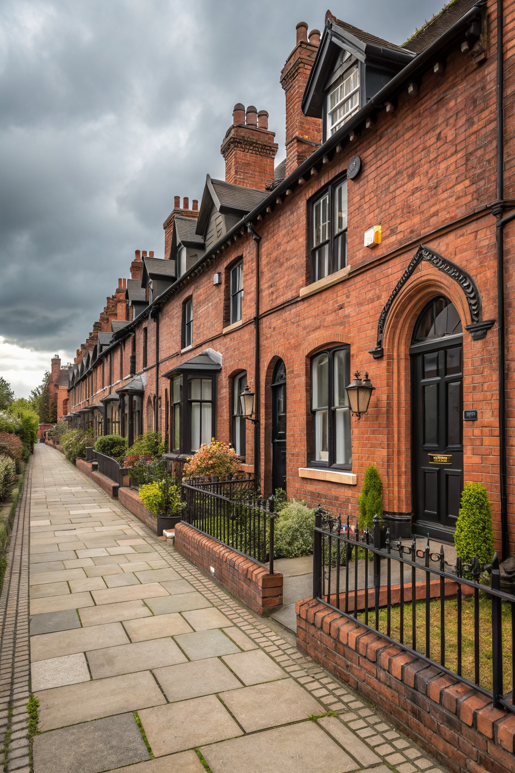 Row of two-story red brick terraced houses with black window frames, doors, and trim, fronted by a paved sidewalk, low iron fences, and small garden plantings under a cloudy sky.