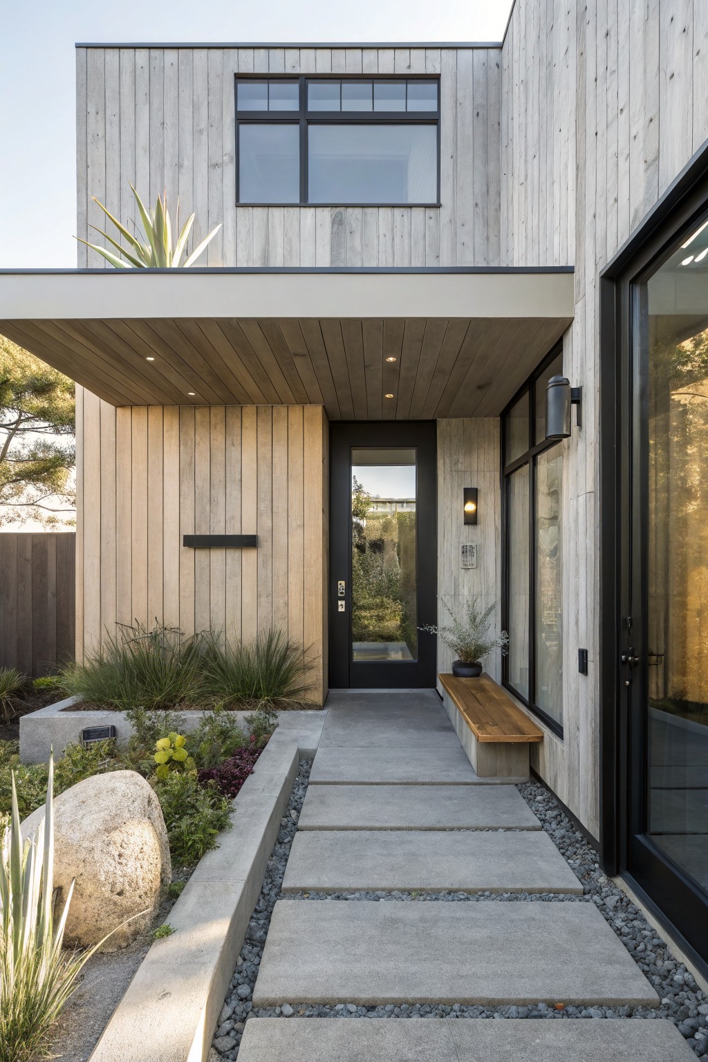 Front entry of a modern house with mixed wood siding, a black door, a wooden bench on a concrete base, and a concrete paver path with gravel and plants.
