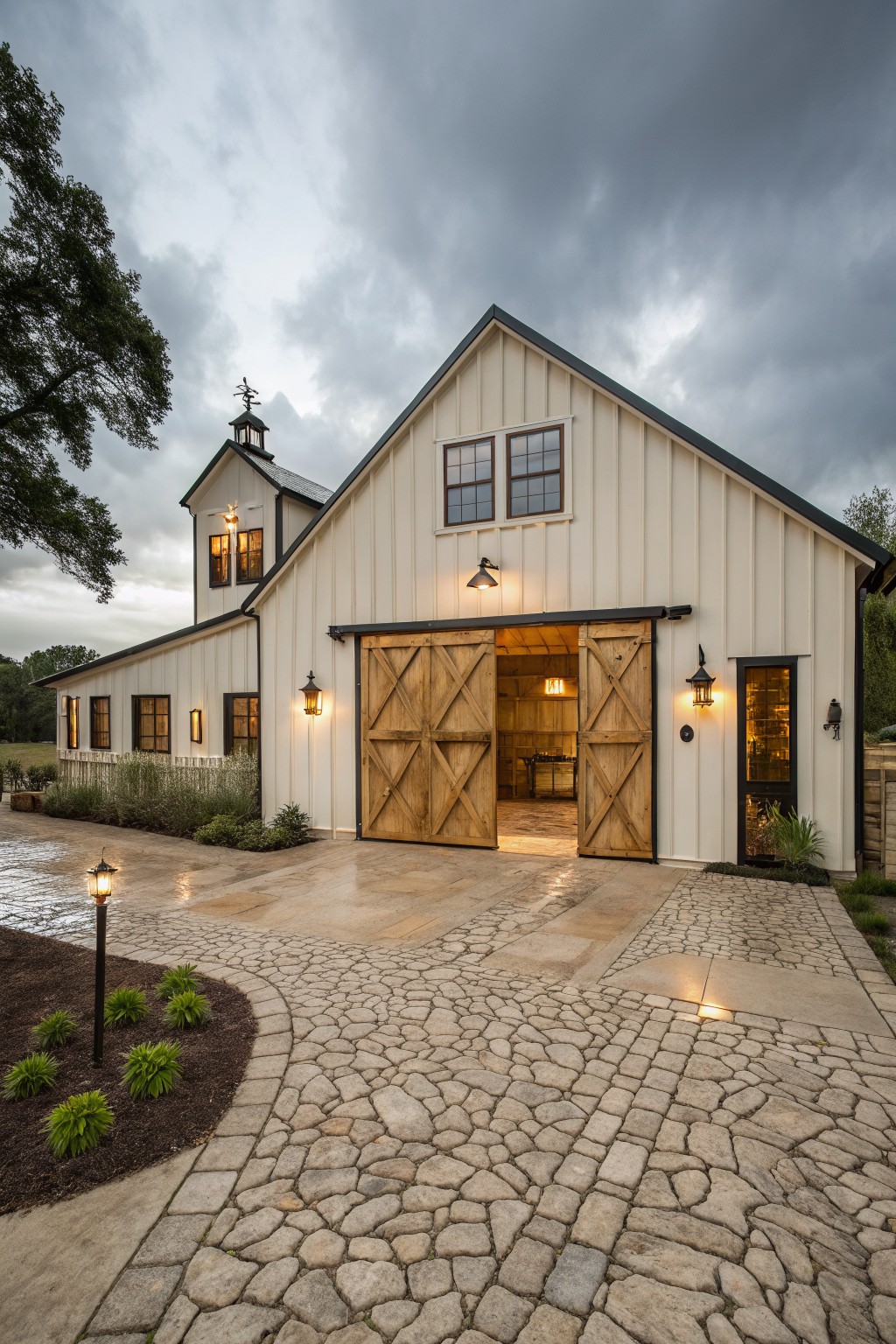 A white barndominium exterior with large wooden barn doors open at the center, a cobblestone driveway, and exterior lanterns under an overcast sky.