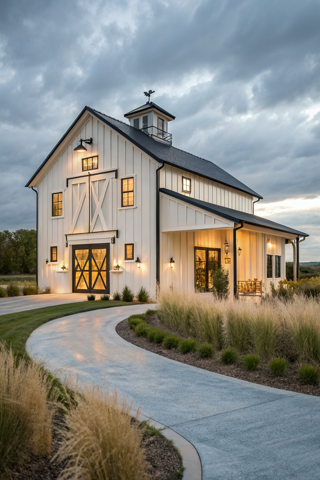 A white barndominium exterior with black trim, large barn-style doors, multiple lit windows, and a small cupola on the roof.
