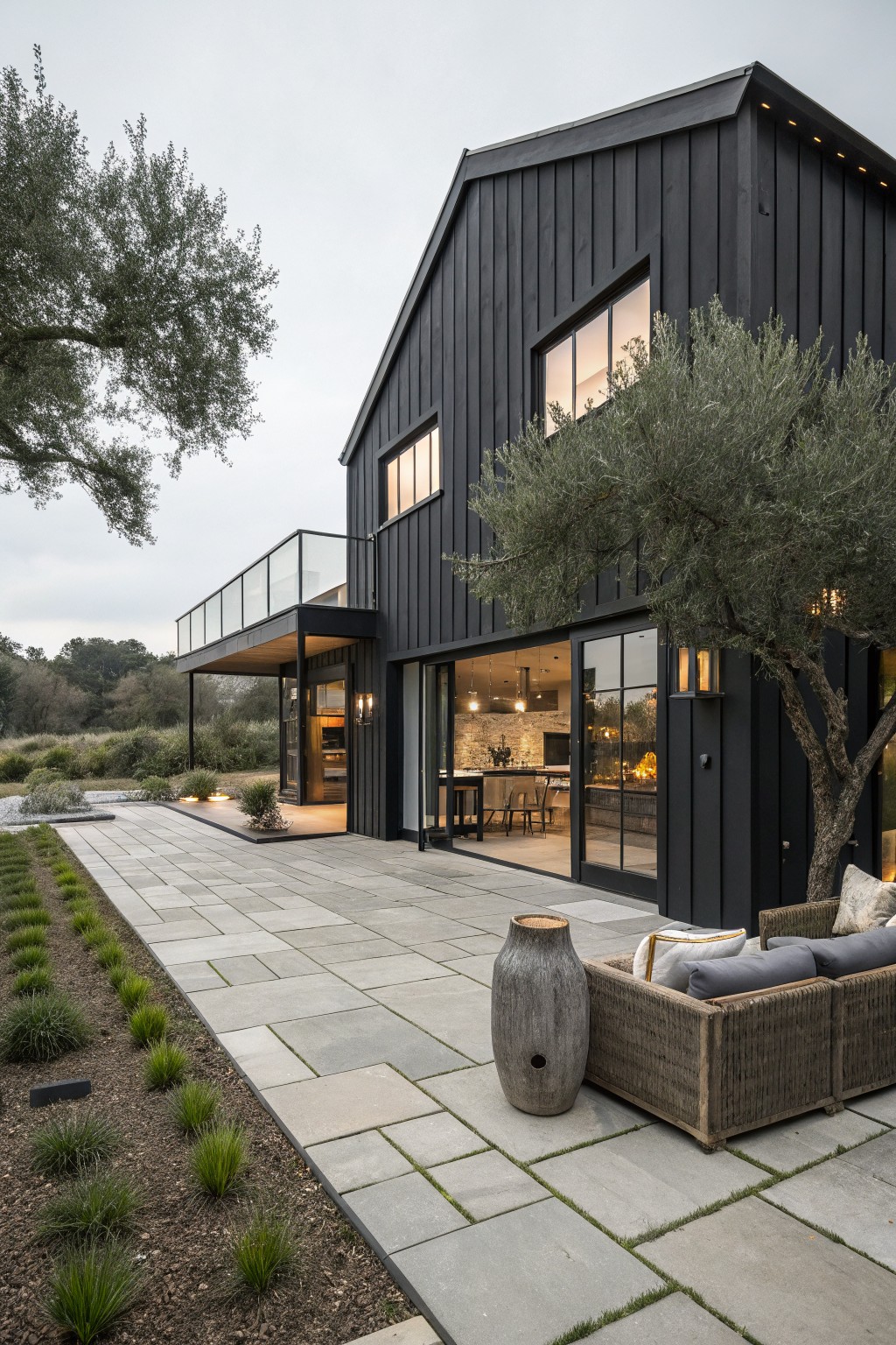 Modern black barndominium exterior with vertical siding, large windows, and open glass doors leading to a stone patio with seating.