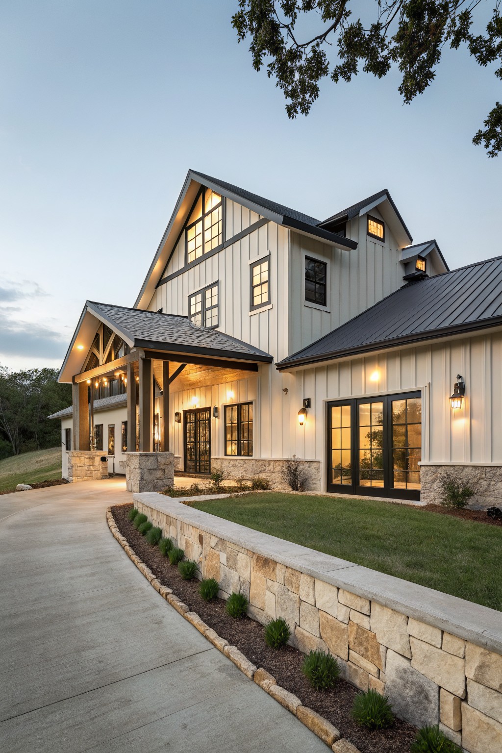 A two-story barndominium exterior with white board and batten siding, dark trim, black-framed windows, and a stone foundation wall along a curved driveway.