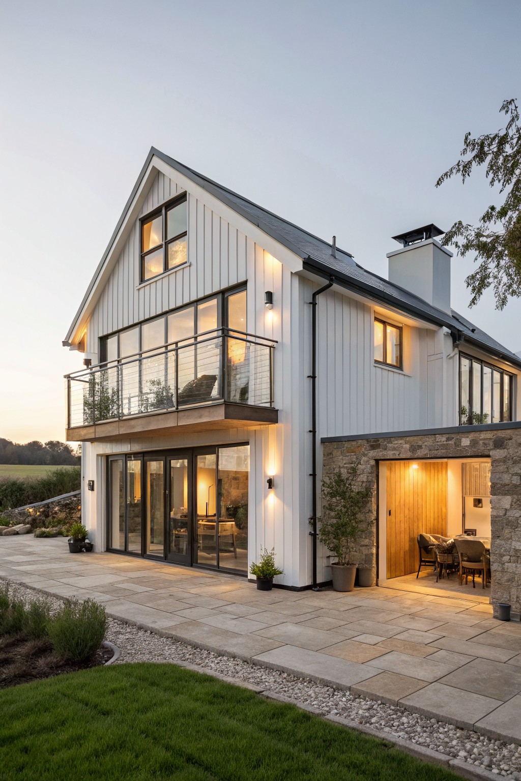 A two-story white barndominium with vertical siding, stone chimney and lower walls, large black-framed windows, a second-floor balcony, and a paved patio area.