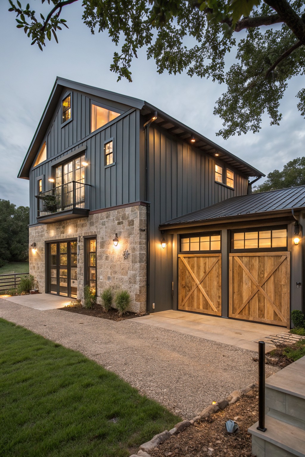 A two-story barndominium exterior at dusk showing dark gray metal siding, a stone lower wall, two wooden barn-style garage doors, and multiple lit windows.