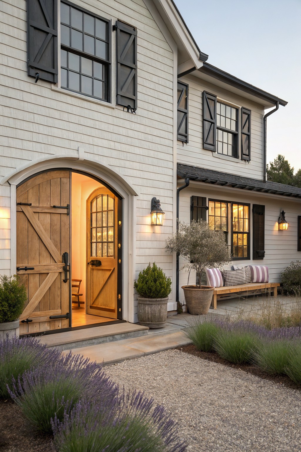 A white farmhouse exterior featuring an arched entry with open wooden double doors revealing an inner door, black shutters on windows, and landscaping with lavender along a gravel path.
