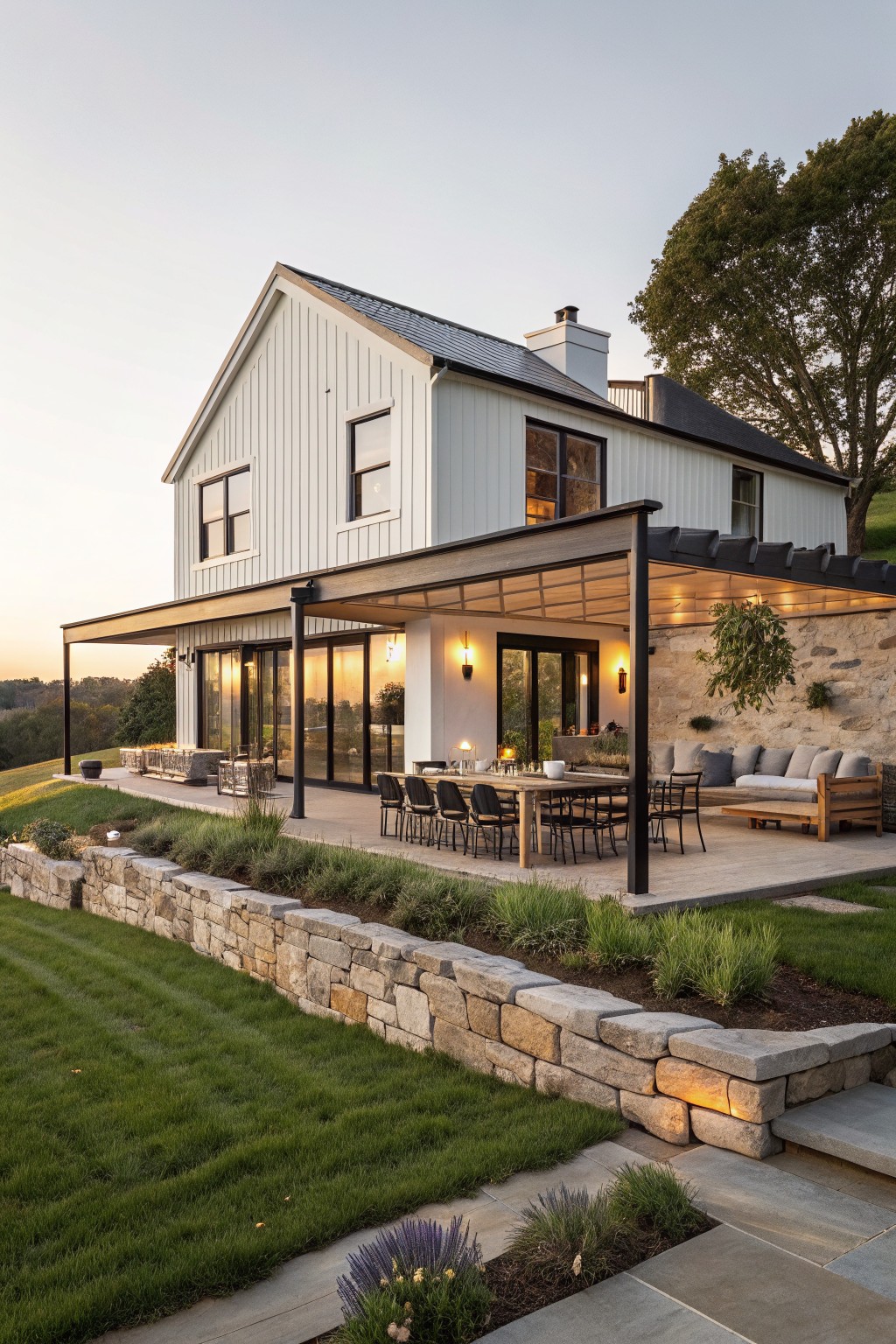 A white modern farmhouse with board and batten siding and a large attached covered patio that includes a dining table, chairs, and seating area next to stone walls and landscaping.
