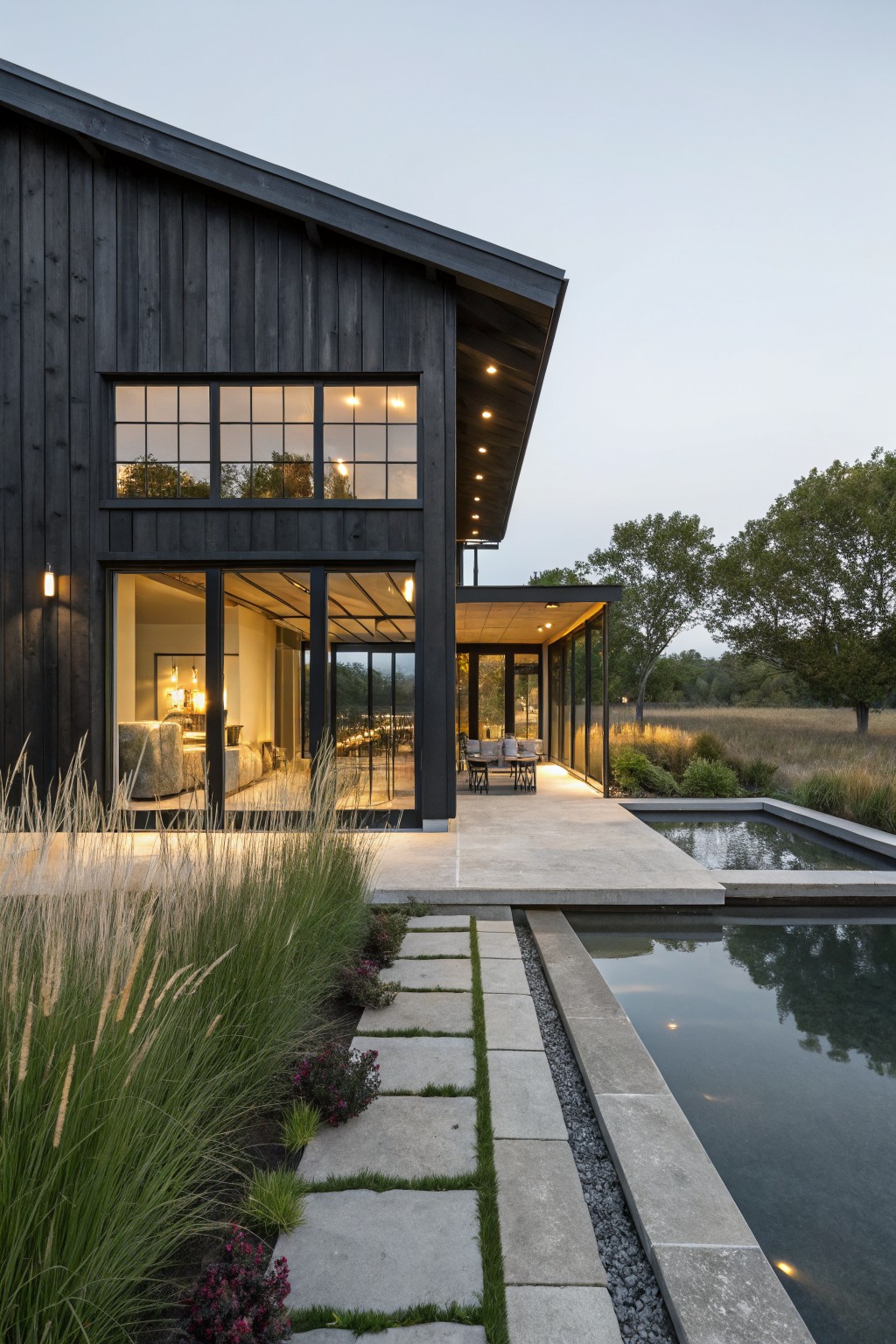 A modern farmhouse exterior with dark wood siding, large black-framed windows, and a concrete patio beside a long reflecting pool.