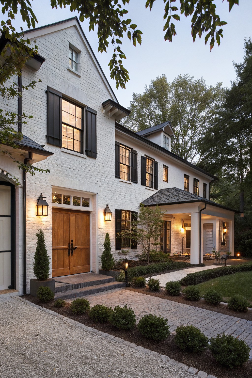 A two-story white brick farmhouse exterior with black shutters, wooden double doors, a covered porch supported by white columns, and landscaped brick pathways at dusk.