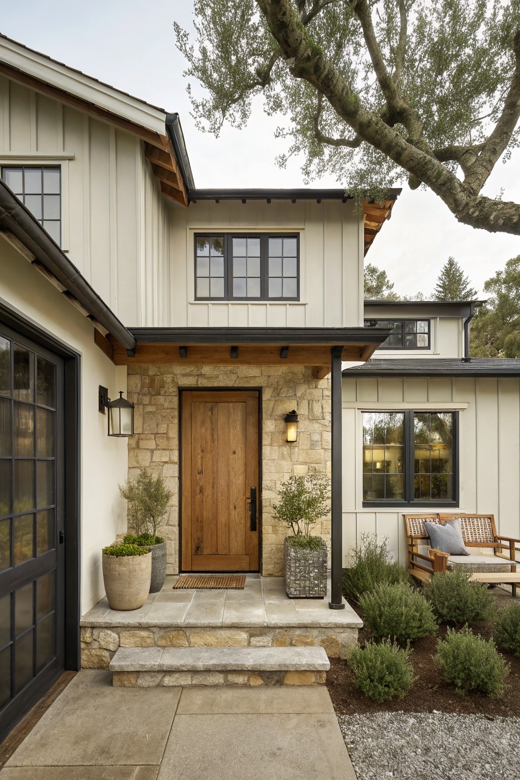 Modern farmhouse entrance with wooden door, stone porch, potted plants, and patio chairs
