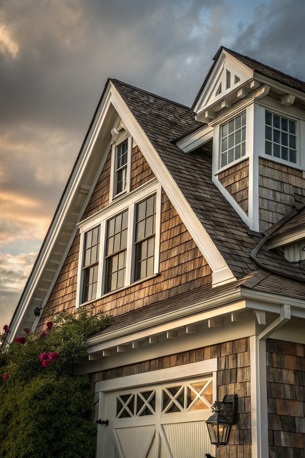 Close-up of a house exterior with brown wood shingle siding, white window trim, dark roof shingles, and a white garage door.