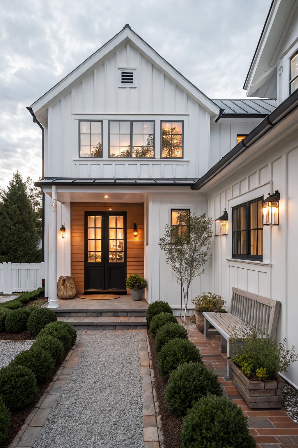 Front view of a small white farmhouse with black-framed windows, a black double door set in wood paneling, stone steps, and a gravel walkway lined with shrubs.